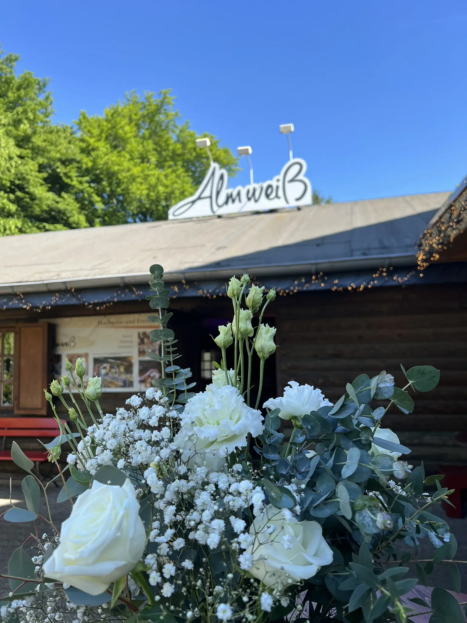 Schriftzug Almweiß auf dem Dach der Blockhütte, davor weiße Rosen