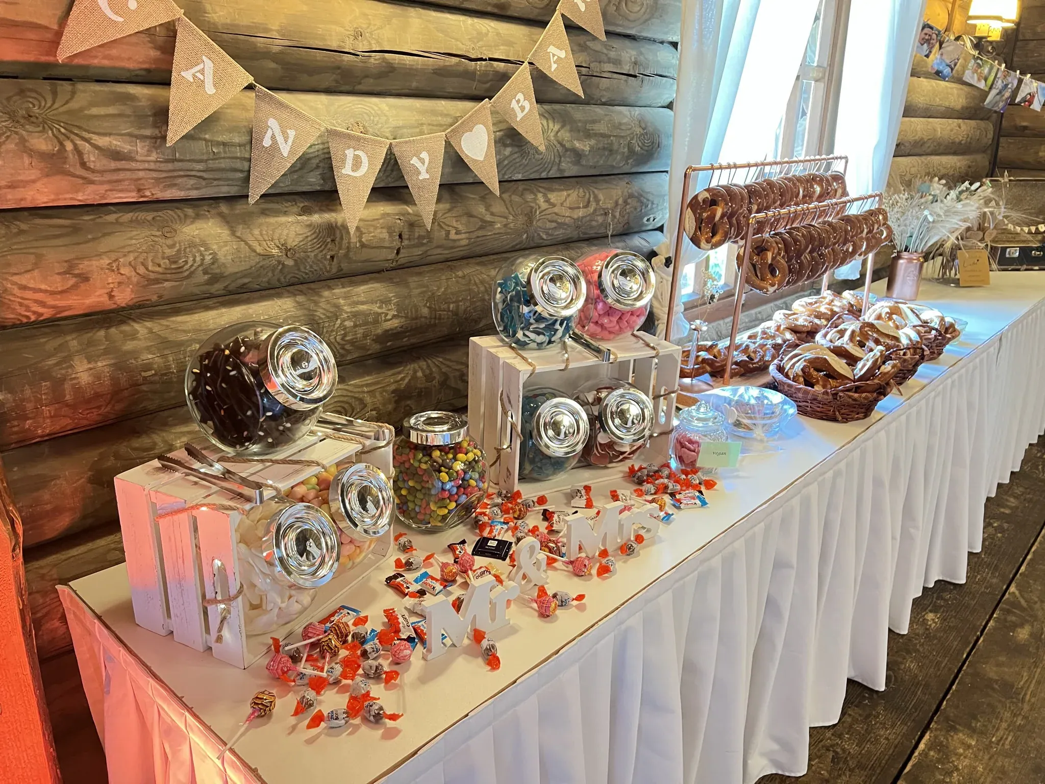 Candybar mit Bonbongläsern, Brezeln und Wimpelkette vor der Blockhauswand