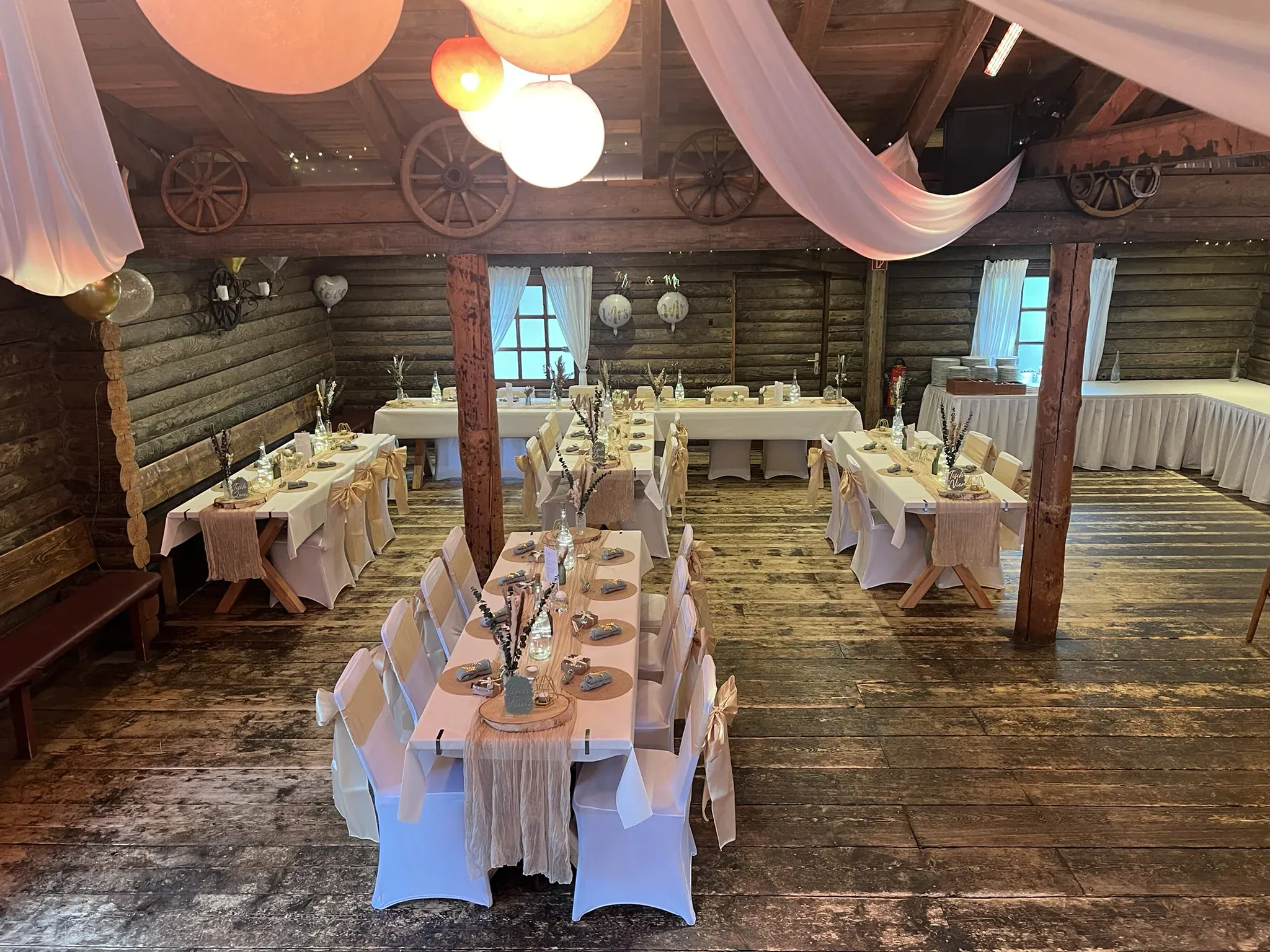 Blockhütte mit Wagenrädern am Balken, Lampions und drei gedeckten Tafeln in Naturtönen